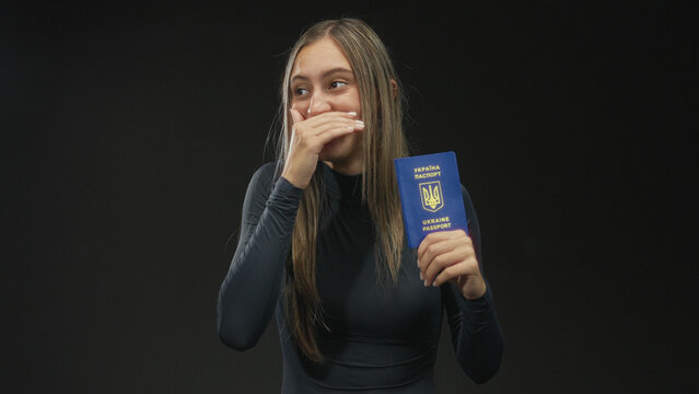 Woman holding ukraine passport with hand covering mouth while laughing and showing passport front in studio black backdrop; joy travel.