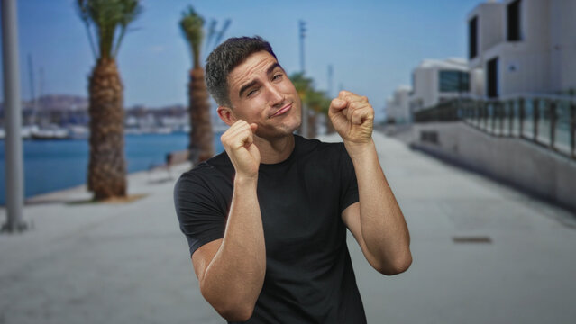 Young hispanic man with fists raised in boxing pose on seaside promenade by palm trees and railings; playful confidence.