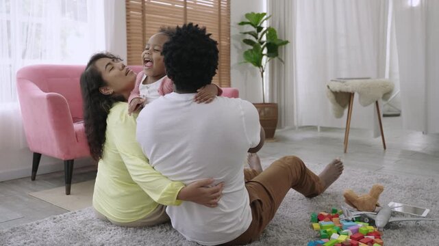 A cute African American child daughter is running and smiling to embrace her father and mother while they sit and wait on the floor, happy and bonding in living room at home. Happy Family Day concept.