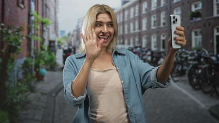 Fototapeta premium Young blonde woman holding smartphone, waving hand and smiling during a selfie call on a narrow european street lined with bicycles and brick buildings; joyful connection.