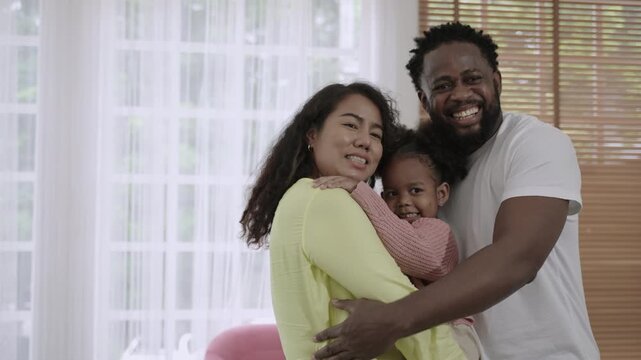 Portrait of a happy African family with father, mother, and their child daughter standing, smiling, and embracing, hugging in love and bonding while looking at the camera in the living room at home. 