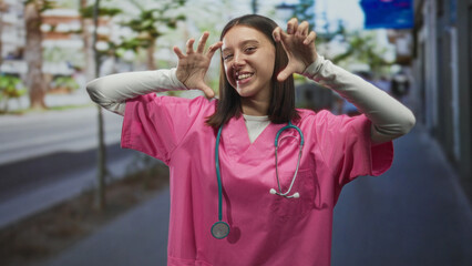 Fototapeta premium Young hispanic nurse woman in pink scrubs with stethoscope makes claw hands framing face on street; playful compassion.