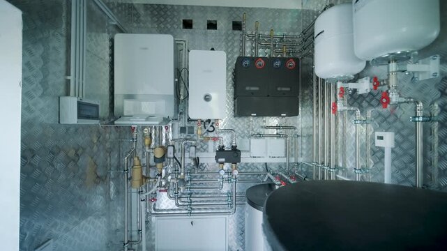 Modern mechanical room interior showing a complex HVAC system with heat pump, boiler, expansion tanks, pipes, and pump groups against a metallic wall.
