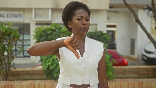 Woman displays thumb down gesture on street in front of storefront wearing white vest and earring; disapproval protest.