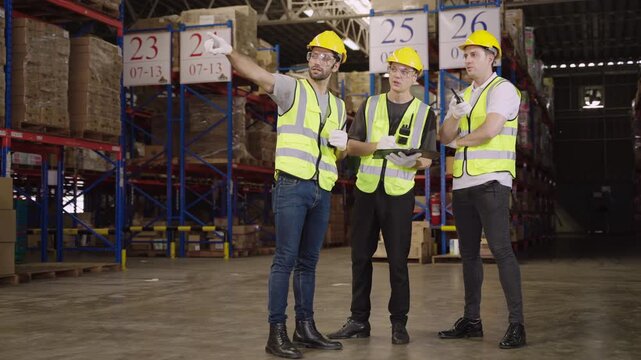 Teamwork of three warehouse workers in uniform wearing yellow safety helmets, standing and working talking and discussing planning in large logistics distribution factory. Inventory stock concept.