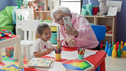 Woman holding smartphone taking selfie with little girl at colorful preschool classroom table showing drawings and colored pencils and toys on shelves  generational bonding joy. © Krakenimages.com