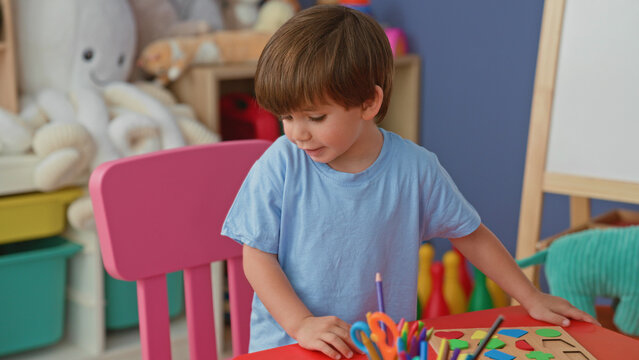 Little boy places wooden shapes into a peg puzzle on a red table inside building; concentration learning.