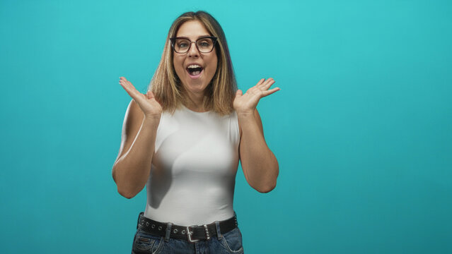 Woman with raised hands and open mouth showing surprise in studio with turquoise backdrop, white sleeveless top and jeans; celebration excitement.