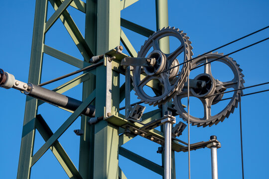 Railway catenary tensioner pulley wheel system on an electric mast under a blue sky in Munich, Germany, showing public transit infrastructure