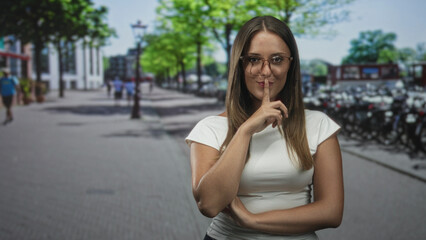 Fototapeta premium Woman with finger to lips shushing on a busy street near parked bicycles and trees, wearing glasses and white top, direct gaze; quiet secrecy.