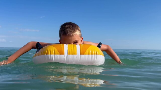 A boy is floating on an inflatable circle in a calm sea under a clear blue sky, enjoying a summer day. The concept includes children's holidays by the sea, safe swimming, summer holidays, and water ac