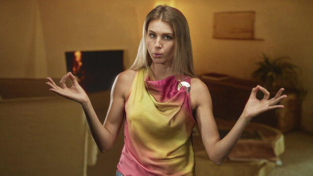 Woman meditating with hands in mudra beside a sofa in a building, wearing colorful sleeveless top and brooch, eyes closed and relaxed; mindfulness calm.
