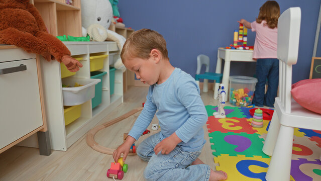 Boy kneeling on floor builds wooden train track and pushes a toy car beside foam puzzle mat and storage shelves with stuffed animals in building; playful childhood joy.