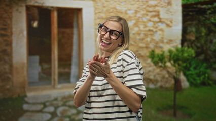 Fototapeta premium Middle age woman wearing black glasses clapping hands by a stone doorway of a rustic building, striped tshirt visible and smiling; joy celebration.