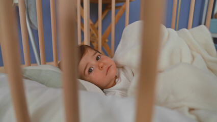 Boy lying in pale wooden crib clutching soft fleece blanket, hand near mouth, gaze sideways toward room window in building  cozy calm. © Krakenimages.com