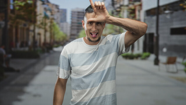 Man sticks out tongue and makes l hand gesture on forehead wearing a striped t shirt on a city street; playful mischief.