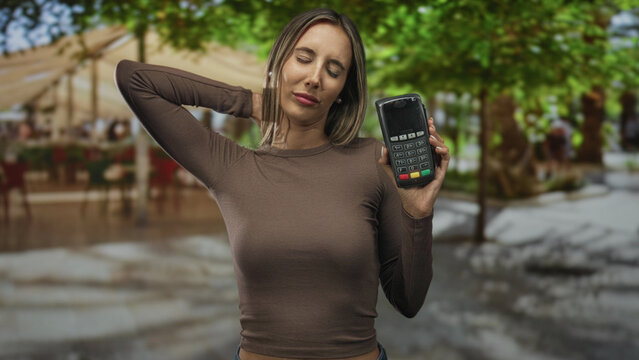 Woman holds card reader with one hand on street cafe terrace, presenting payment terminal to camera; contactless payment confidence.