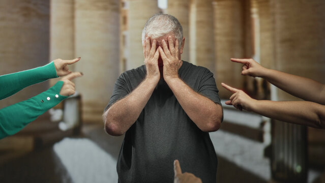 Senior man with hand covering face and multiple fingers pointing at him in a building corridor; humiliation.