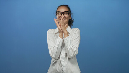 Fototapeta premium Woman clapping hands and smiling in a blue studio, wearing glasses and a white shirt; joy celebration.