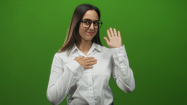 Woman with hand on chest and raised hand waving, smiling while wearing white shirt and glasses in studio; warm friendly greeting.