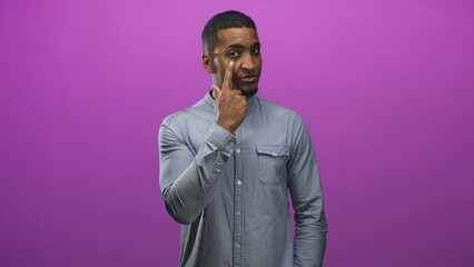 Fototapeta premium Young black man points finger to eye wearing a blue button shirt in a purple studio backdrop; thoughtful curiosity.