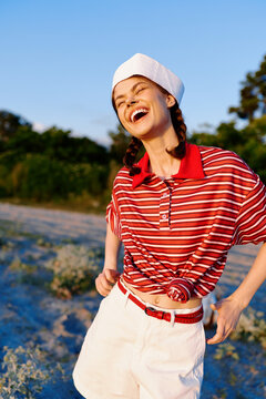 A young woman on a sunny beach laughs in a red striped shirt and white shorts, embodying carefree summer style and outdoor vitality.