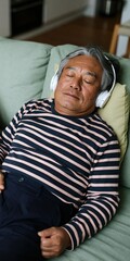 Fototapeta premium Older man resting on couch with headphones and striped shirt eyes closed for relaxation background and sleep therapy design
