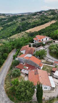Vertical aerial drone view of rural houses with orange roofs by winding countryside road surrounded by green hills and trees in Italy region landscape.