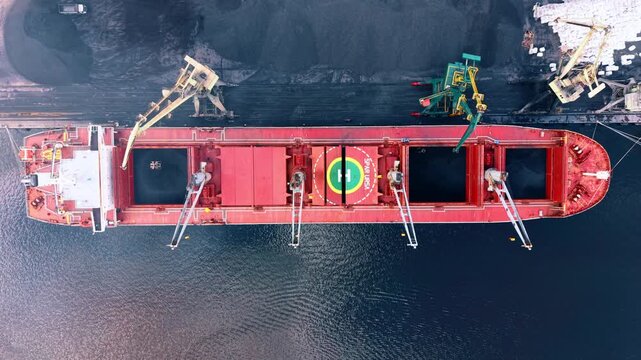 Red bulker for transportation of unpackaged raw commodities. Top view on the vessel during the process of coal unloading.