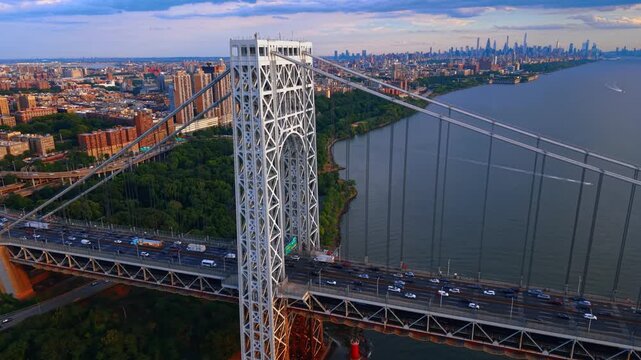 Flight along the George Washington Bridge at sunset. Multiple transport moves by the bridge. The Washington Heights neighborhood in Upper Manhattan, New York City in the backdrop.