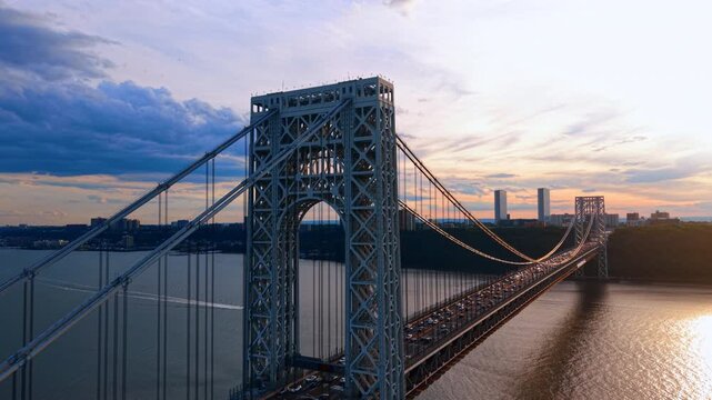 Heading to the top of the massive metal support of the George Washington Bridge at sunset. Lots of traffic on the structure. Boat is moving by the river.