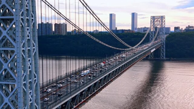 Numerous cars move in the hectic traffic by the George Washington Bridge. Green waterfront in the backdrop at sunset time.