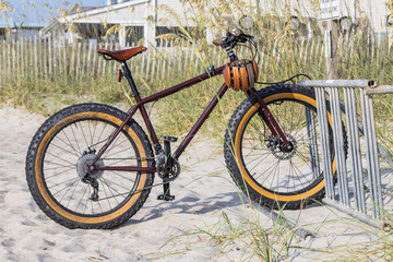 Fototapeta premium Sturdy bicycle rests on sandy beach beside wooden fence. Thick tires suited for rough terrain. Rustic brown frame and seat contrast with golden wheels. Natural light highlights texture of sand