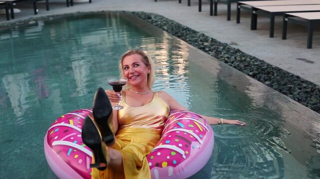 A blonde woman in a yellow swimsuit floats on a giant pink donut inflatable ring in a turquoise pool, relaxing with one hand raised to her face in a playful pose