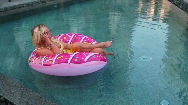 A blonde woman in a yellow swimsuit floats on a giant pink donut inflatable ring in a turquoise pool, relaxing with one hand raised to her face in a playful pose