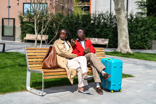 Thoughtful african american couple man and woman tourists relaxing on bench near hotel while waiting for taxi, positive black male and female travelers resting together and awaiting drive to airport.