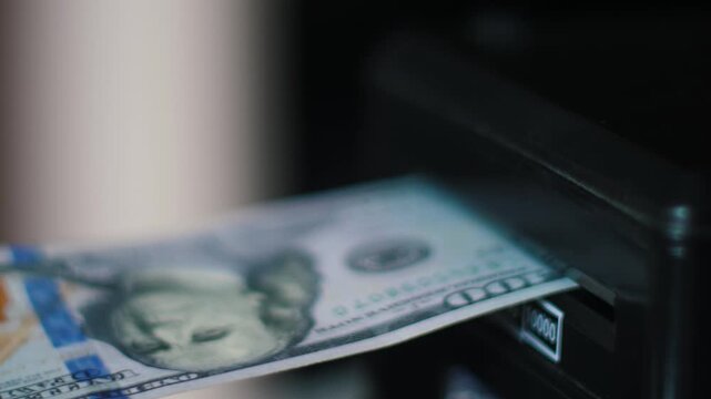 A close-up of a black automatic device into whose slot one-hundred-dollar bills are alternately drawn. American banknotes quickly disappear into the bill acceptor of a terminal 