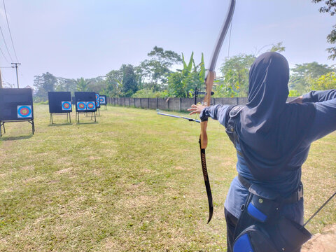 Archery enthusiast focuses intently on target. Grassy field hosts multiple shooting stations. Clear skies enhance outdoor training atmosphere. Person wears hooded gear and draws bow with precision