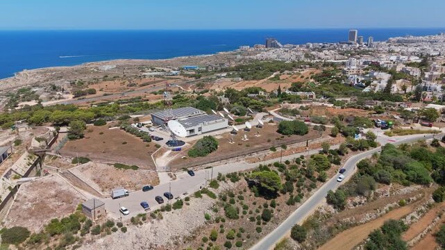 Madliena, Malta satellite communication ground station with parabolic antenna arrays, telecom infrastructure, solar panels and utility buildings at an aerial outdoor facility.