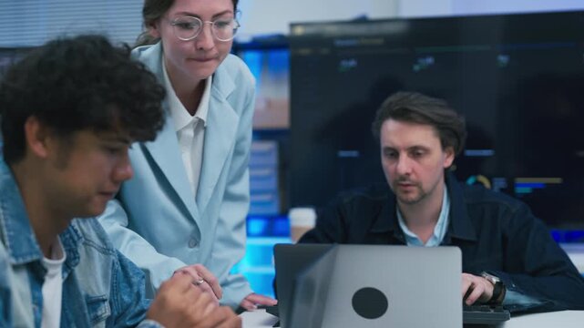 collaborative AI development team in modern tech office: young male developer coding on laptop while senior colleague leans over discussing code and data visualizations on multiple monitors	
