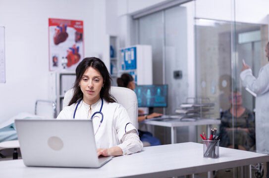 Professional doctor in white coat working on laptop in modern hospital office room, therapist typing on computer consult patient online, makes research, analyzing results information from internet.