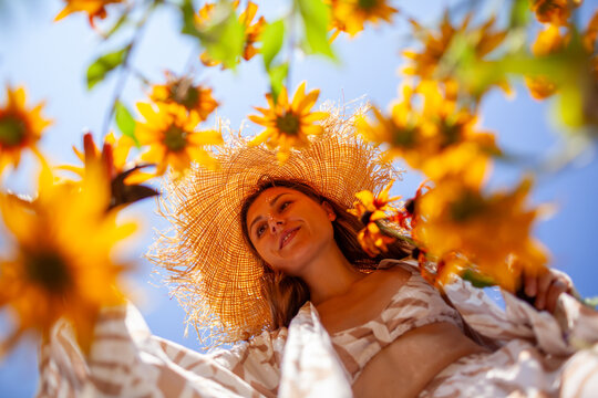 Woman picking rudbeckia flowers using pruner in summer garden. Portrait from below. Stylish gardener wearing straw hat against sky