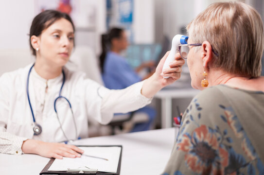 Medic uses a digital thermometer gun to check a senior patient temperature during a clinical examination and health screening in a hospital office.