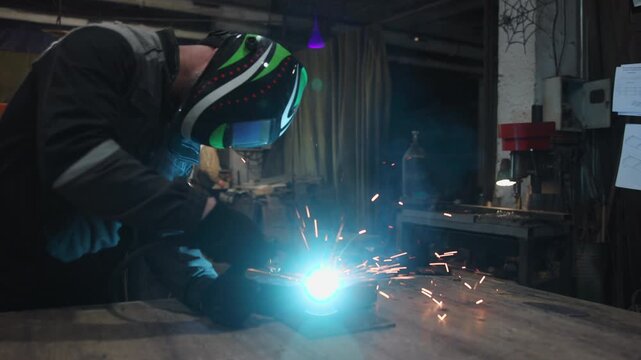 Helmeted welder creating bright blue arc and orange sparks over metal workpiece in dim industrial workshop beside cluttered bench and focused secondary worker, with welding, welder, helmet, spark