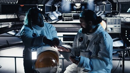 An astronaut reviews onboard analytics on a tablet while talking to a colleague  collaborating cosmonauts track star and asteroid patterns during a deep space survival mission and scientific research. © DC Studio