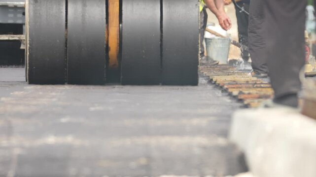 Black road roller presses the pavement surface. Worker in dark pants walks beside the machine. Concrete blocks mark the road's side. Truck stands in the distance