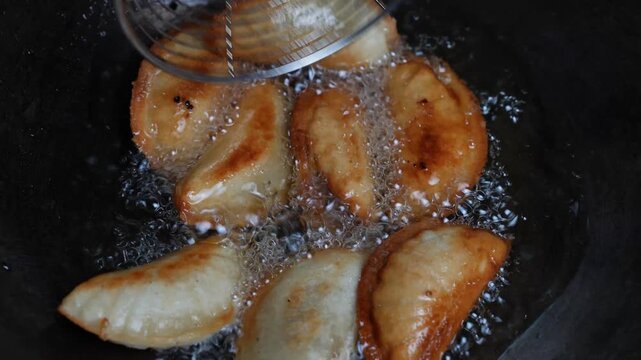 Fried dumplings cooking in hot oil in a dark pan, with bubbles and golden browned crusts close up