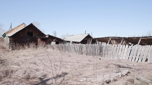 Abandoned rural homestead with leaning picket fence and derelict wooden barns under clear pale blue sky