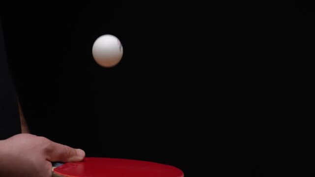 This single keyframe captures a ping pong ball mid-air above a red paddle. Hand grips the paddle steadily during play. Black background emphasizes motion and focus