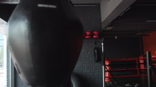 Close-up heavy punching bag inside industrial boxing gym with red padded ring corner, mirrored wall, hanging speed bag, stacked ropes, support poles, and strong indoor contrast